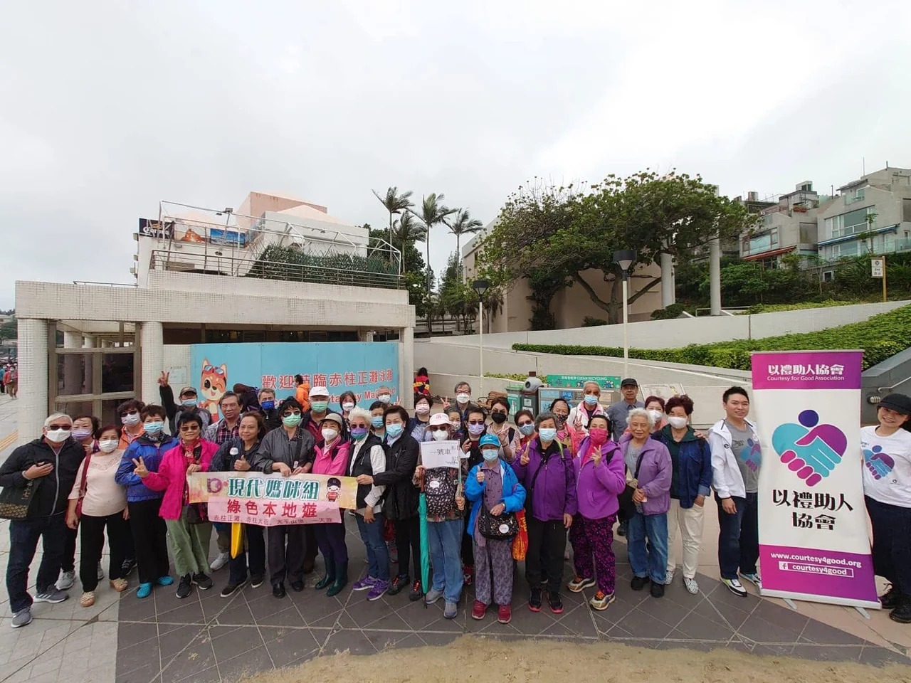 Older participants and volunteers posing outdoors, with Stanley Main Beach signage and the Association's pull-up banner in the background.